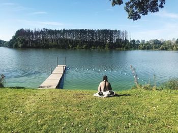 Rear view of man sitting on grass by lake against sky