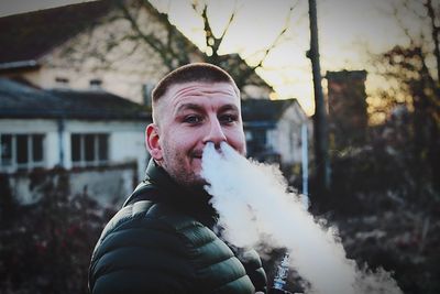 Portrait of young man emitting smoke from nose while standing against house during sunset