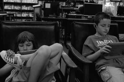 Siblings using digital tablets while sitting on chairs in library
