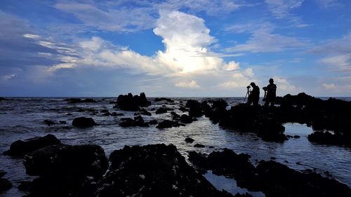 Silhouette people standing on rock in sea against sky