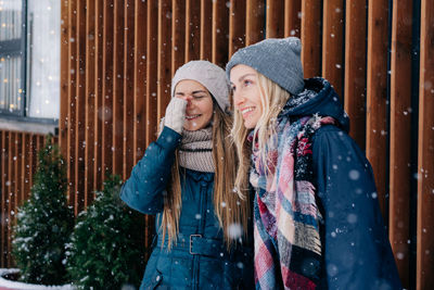 Attractive young women are laughing while standing under the snowfall in warm winter clothes.