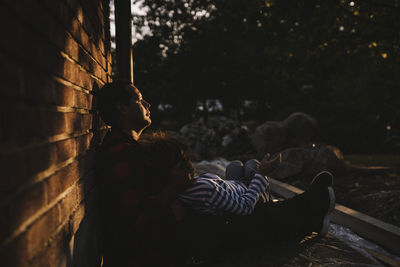 Side view of father and son resting by brick wall during sunset