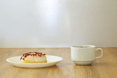 Close-up of donut in plate with coffee cup on wooden table against white wall