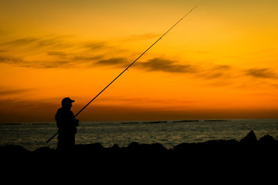 Silhouette man fishing in sea against sunset sky