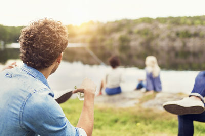 Men having drink while female friends fishing at lakeshore