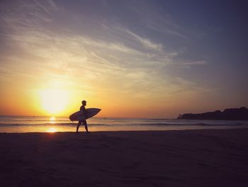Man with surfboard walking on seashore during sunset