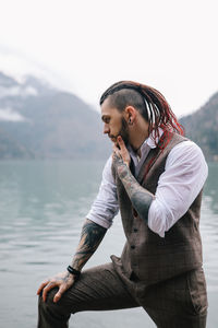 A man a groom a hipster with dreadlocks in a wedding suit stands by sea and mountains in nature