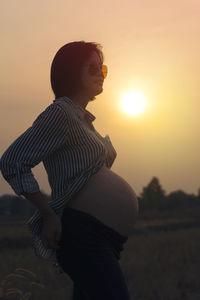 Pregnant woman wearing sunglasses standing against sky during sunset