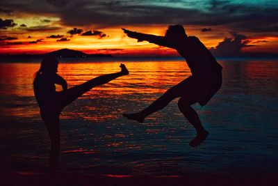 Silhouette of woman kicking man while standing at beach against sky during sunset