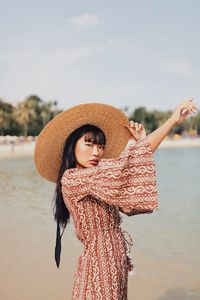 Portrait of young woman wearing hat standing at beach against sky