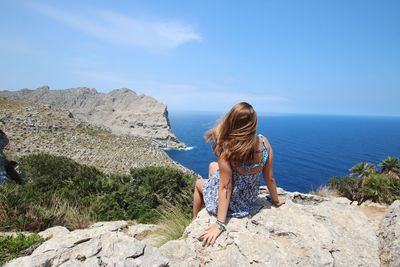 Rear view of woman sitting on rock by sea against sky