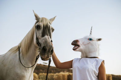 Young woman wearing unicorn mask gently stroking white horse