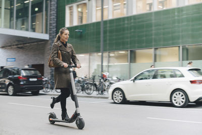 Full length of confident businesswoman riding electric push scooter on street in city