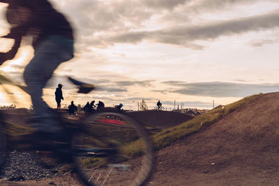 People riding bicycle on hill against sky during sunset