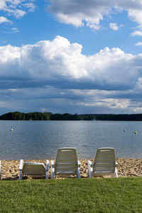 Deck chairs by lake against cloudy sky