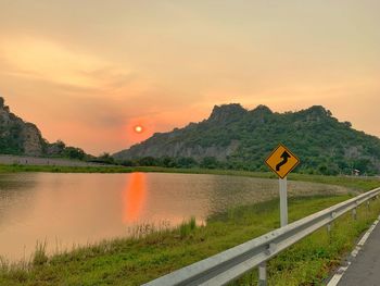 Road sign by lake against sky during sunset