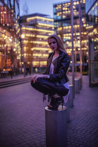 Portrait of young woman crouching on bollard in city at night