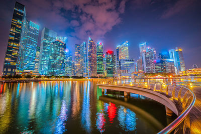 Illuminated modern buildings by river against sky in city