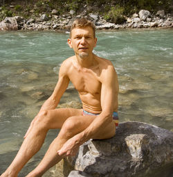 Portrait of shirtless man sitting on rock against river