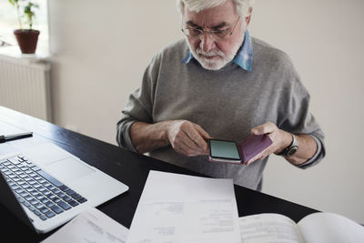 Senior man with laptop holding mobile phone while reading bills at table