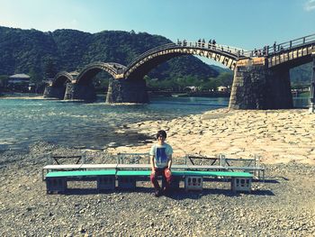 Portrait of young man sitting on bench against arch bridge