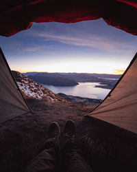 Low section of person in tent by lake against sky during sunset
