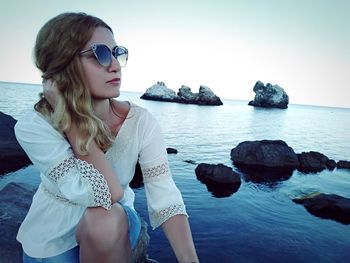 Woman wearing sunglasses while sitting on rock in sea against sky