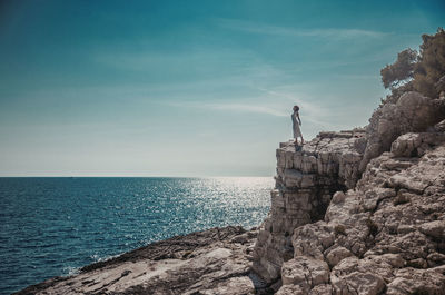Teenage girl standing on cliff by sea against sky