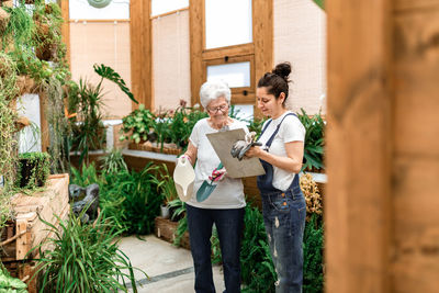 Adult woman writing on clipboard near elderly colleague while working in hothouse together