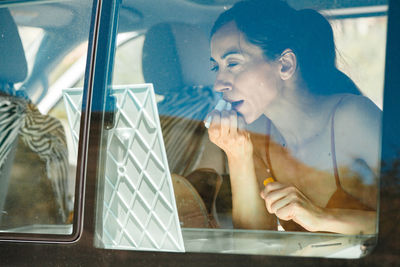 Woman applying make-up in front of mirror in car seen through window