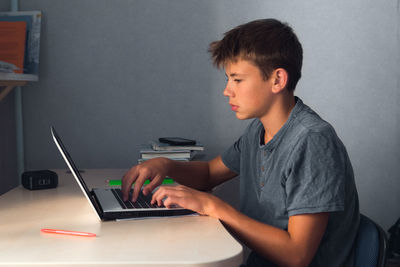 Student teenager boy doing homework with laptop, open copybook and computer, workplace at home
