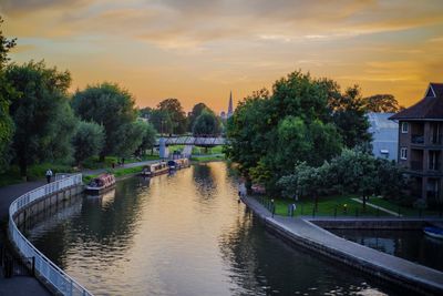 Bridge over river amidst trees and buildings against sky during sunset