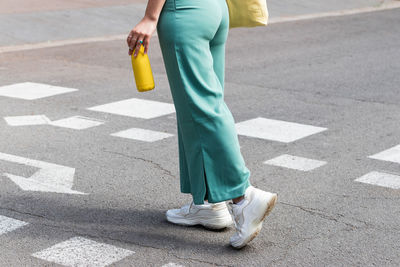 Side view of crop anonymous female with bottle of water walking along crosswalk in city