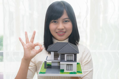 Portrait of young woman with model house gesturing ok sign