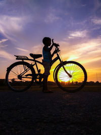 Silhouette boy riding bicycle against sky during sunset