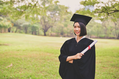 Smiling young woman in graduation gown holding certificate while standing at park