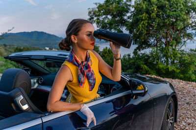 Young woman sitting in a cabriolet and looking at the sunset through binoculars