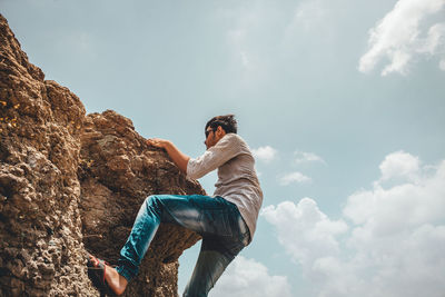 Low angle view of young man clambering on rock against sky during sunny day