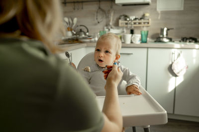 Mother feeding baby girl with porridge in kitchen