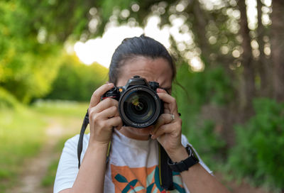 Front view of female photographer standing outdoors, holding camera and pointing it toward viewer