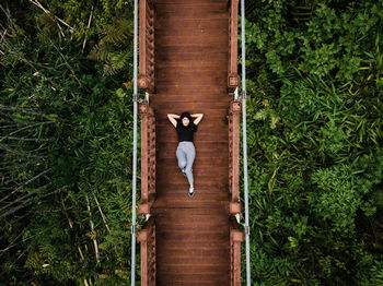 Directly above portrait of woman lying on footbridge over trees in forest