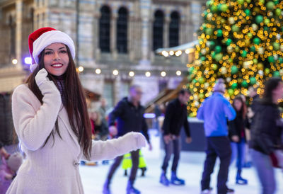 Smiling young woman standing by christmas tree in city during winter