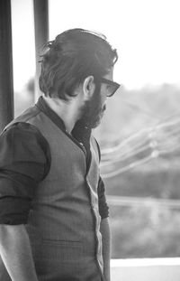Side view of young man looking away while standing against window