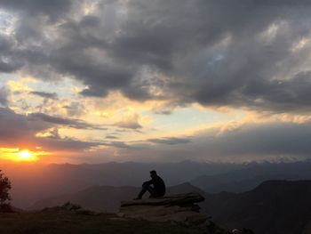 Silhouette man sitting on rock by mountains against cloudy sky during sunset