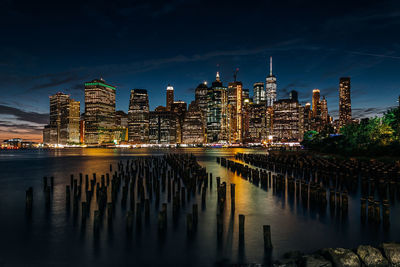Illuminated modern buildings by river at manhattan in city during night