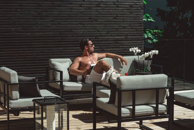 Shirtless man wearing sunglasses sitting on sofa against wall