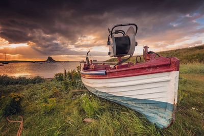 Boat moored on grassy shore against cloudy sky during sunset