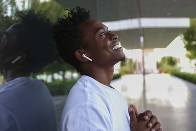 Smiling man with wireless in-ear headphones day dreaming leaning on wall