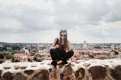 Portrait of young woman sitting on retaining wall against cityscape