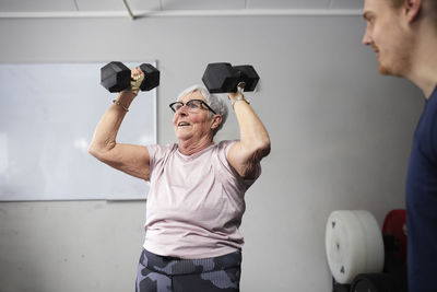 Senior woman exercising with dumbbells by male fitness instructor in gym